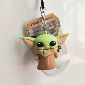 Może przedstawiać: Zielony i brązowy brelok Baby Yoda ze srebrnym metalowym zapięciem. Figurka ma duże czarne oczy i duże uszy. Na etykiecie widnieje napis "TRAVEL DANGLE".