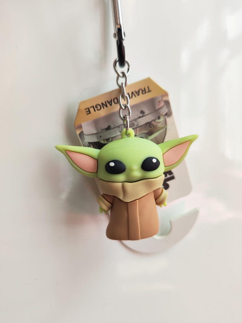 Może przedstawiać: Zielony i brązowy brelok Baby Yoda ze srebrnym metalowym zapięciem. Figurka ma duże czarne oczy i duże uszy. Na etykiecie widnieje napis "TRAVEL DANGLE".