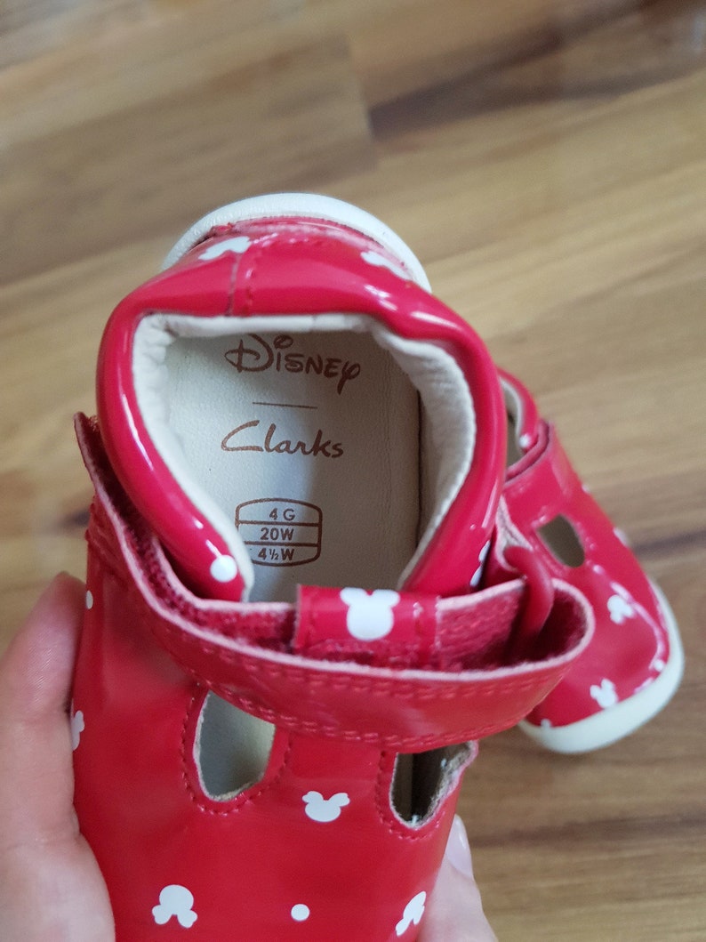 Pu&ograve; includere: Sandali rossi in vernice con stampa bianca di Topolino. I sandali hanno una chiusura a velcro e una suola bianca. L'interno del sandalo ha un'etichetta che recita "Disney Clarks" e "4G 20W 4 1/2 W".