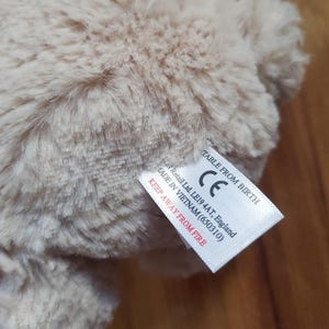 May include: A close-up of a beige plush toy with soft, textured fur. A white tag is attached, with text including "TABLE FROM BIRTH" and "KEEP AWAY FROM FIRE".