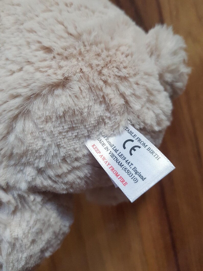 May include: A close-up of a beige plush toy with soft, textured fur. A white tag is attached, with text including "TABLE FROM BIRTH" and "KEEP AWAY FROM FIRE".