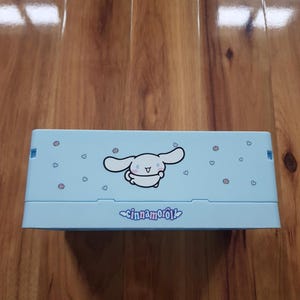 Puede incluir: Un estuche azul claro con un personaje de dibujos animados y el texto "cinnamonroll". El estuche está decorado con pequeños corazones rosas y tiene forma rectangular.