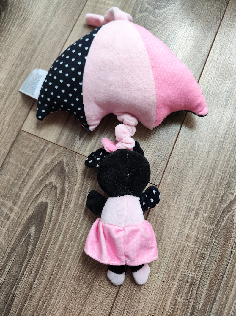 K&ouml;nnte beinhalten: Ein schwarzes und pinkfarbenes Pl&uuml;schtier mit einem rosa Regenschirm. Das Spielzeug hat einen schwarzen Kopf mit wei&szlig;en Punkten und einen rosa Rock mit wei&szlig;en Punkten.