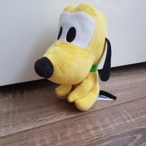 Pode incluir: Um cão de pelúcia amarelo com orelhas pretas e uma coleira verde. O cão tem olhos grandes e brancos com pupilas pretas.