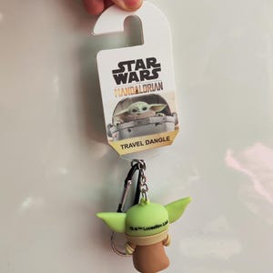 Może przedstawiać: Star Wars Mandalorian brelok podr&oacute;żny z Grogu, znanym r&oacute;wnież jako Baby Yoda. Brelok ma zieloną figurkę Grogu z dużymi uszami, brązową szatą i srebrnym k&oacute;łkiem na klucze. Na opakowaniu widnieje napis "Star Wars Mandalorian" i "Travel Dangle."