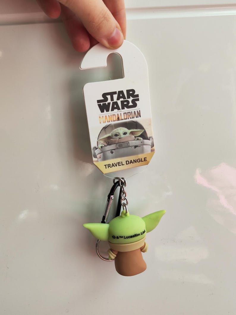 Może przedstawiać: Star Wars Mandalorian brelok podr&oacute;żny z Grogu, znanym r&oacute;wnież jako Baby Yoda. Brelok ma zieloną figurkę Grogu z dużymi uszami, brązową szatą i srebrnym k&oacute;łkiem na klucze. Na opakowaniu widnieje napis "Star Wars Mandalorian" i "Travel Dangle."
