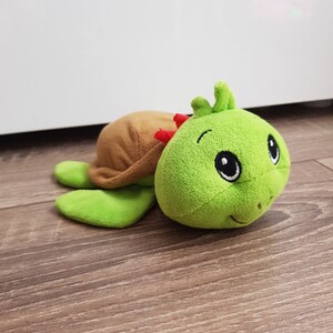 May include: A green plush turtle toy with a brown shell and a smiling face. The toy has large black eyes and a red nose.