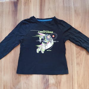 Puede incluir: Camiseta negra de manga larga con un gráfico de Buzz Lightyear de Lightyear de Disney Pixar. El diseño muestra al personaje en una pose de vuelo con el logotipo de la película. La camiseta tiene cuello redondo con ribete azul.