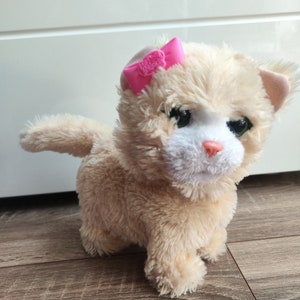 Op de afbeelding: Een pluche kat met een roze strik op zijn kop. De kat is lichtbruin met witte vacht op zijn gezicht en borst. Hij heeft groene ogen en een roze neus.