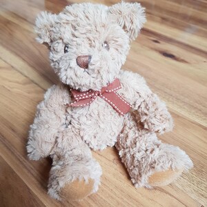 May include: A soft teddy bear with light brown fur and a brown nose. The bear has a red bow with white polka dots. The bear is sitting on a wooden surface.