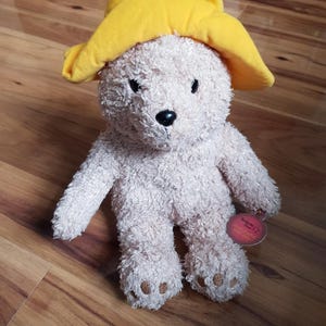 Può includere: Un orsacchiotto di peluche beige con un cappello giallo. L'orso ha occhi marroni e un naso nero. L'orso è seduto su un pavimento di legno. L'orso ha un'etichetta che dice "Cuddle Bear".