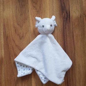 May include: A white elephant-shaped baby security blanket. The elephant head is plush with small ears and a trunk. The blanket portion is white with a gray star pattern on the underside of the corners. A soft, comforting item for infants.