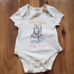 Może przedstawiać: Kremowe body dla niemowląt z grafiką Daisy Duck i napisami "Hello Sunshine!" oraz "Daisy Duck". Body z krótkim rękawem ma zatrzaski na dole i jest wykonane z miękkiej tkaniny.