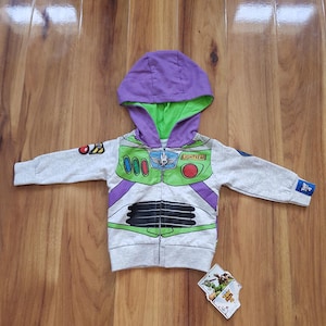 Puede incluir: Una sudadera con capucha gris con cremallera, capucha morada y forro verde, diseñada para parecerse al traje espacial de Buzz Lightyear. La parte delantera presenta un panel verde en el pecho con el número "000" y la palabra "LIGHTYEAR". Una pequeña etiqueta con una imagen de dibujos animados está en la parte inferior.