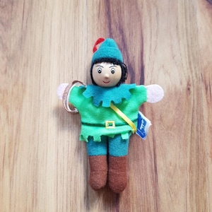 Könnte beinhalten: Ein grünes und braunes Plüschspielzeug von Peter Pan mit einem grünen Hut und einem gelben Gürtel. Das Spielzeug hat ein Holzgesicht mit bemalten Gesichtszügen und hält einen Holzring.