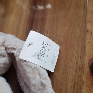 May include: A beige bunny toy with long ears, with a tag that reads "next BABY next.co.uk". The toy is made of a soft, fluffy material.
