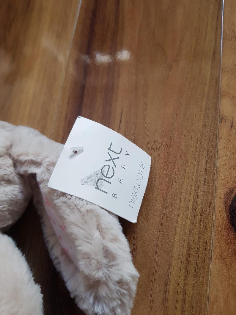 May include: A beige bunny toy with long ears, with a tag that reads "next BABY next.co.uk". The toy is made of a soft, fluffy material.