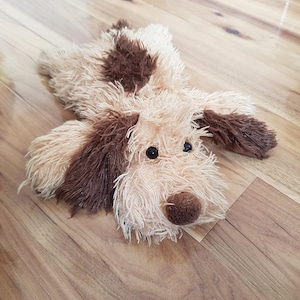 May include: A plush dog toy featuring shaggy, cream-coloured fur with dark brown patches on the back and ears. The stuffed animal has two black plastic eyes and a round, dark brown fabric nose, designed in a lying-down position with floppy ears.
