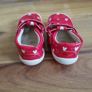 Pu&ograve; includere: Un paio di scarpe rosse per bambini piccoli con orecchie di Topolino bianche stampate su di esse. Le scarpe hanno cinturini bianchi e suole bianche.