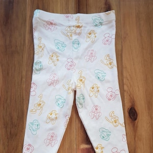 May include: White leggings featuring a repeating pattern of cartoon characters in pink, yellow, teal, and orange outlines. The characters include Mickey Mouse, Donald Duck, and Daisy Duck. The leggings are made of a soft, stretchy fabric.