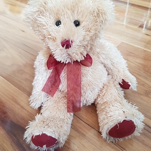 Könnte beinhalten: Ein Plüschteddybär mit hellbraunem Fell, schwarzen Augen und einer weinroten Schleife. Die Pfoten und die Nase des Bären sind ebenfalls weinrot. Der Bär sitzt auf einer Holzoberfläche.