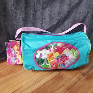 Puede incluir: Una bolsa de deporte turquesa con una correa rosa y un gráfico colorido de personajes de Trolls. Una caja a juego con la palabra "Trolls" está al lado de la bolsa. La bolsa tiene cierre de cremallera.