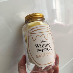 Può includere: Un barattolo di plastica trasparente con coperchio dorato, pieno di spugnette per il trucco gialle. Il barattolo ha un'etichetta con il logo Disney Winnie the Pooh e le parole "MAKEUP SPONGE" in testo marrone.