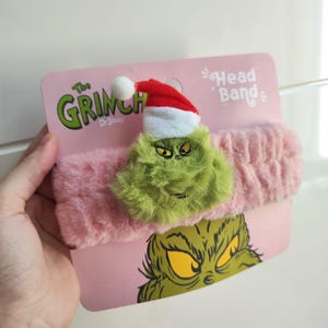 Op de afbeelding: Een roze haarband met een groene Grinch-figuur met een kerstmuts. De haarband wordt getoond op een roze kaart met de woorden "The Grinch" en "Head Band" erop.