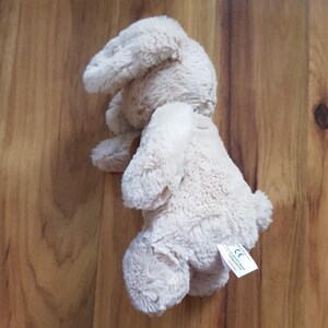 May include: A light beige plush bunny rabbit toy. The stuffed animal has long, floppy ears and a small, fluffy tail. The toy is lying on a wooden surface. A small tag is attached to the toy.