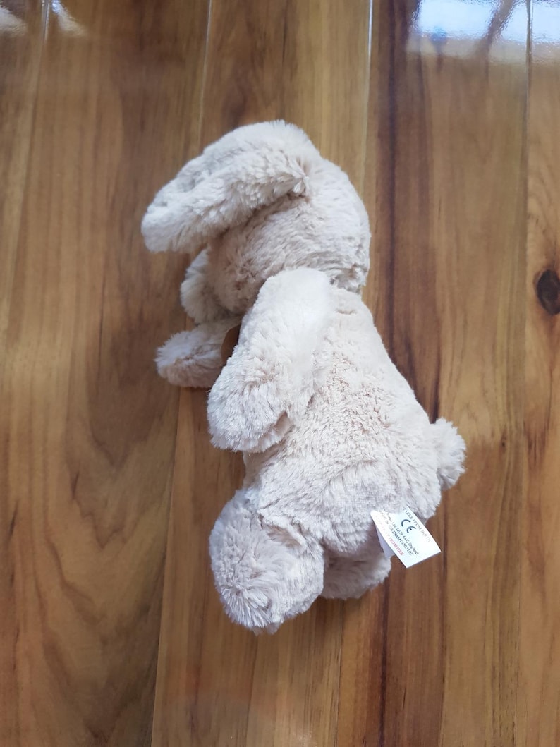 May include: A light beige plush bunny rabbit toy. The stuffed animal has long, floppy ears and a small, fluffy tail. The toy is lying on a wooden surface. A small tag is attached to the toy.
