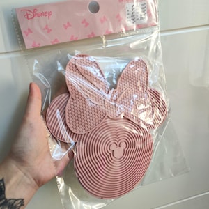 Peut inclure: Un nettoyant pour pinceaux de maquillage rose en forme de Minnie Mouse dans un emballage plastique transparent. Le nettoyant a un nœud et un motif circulaire avec le logo Disney sur l'emballage. L'emballage comprend également des informations sur les tailles.