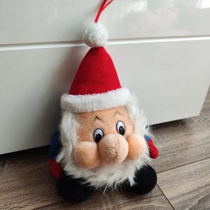 Op de afbeelding: Een pluche kerstversiering van de Kerstman. De versiering heeft een rode muts met een witte pompon en rand, een witte baard en een lachend gezicht. De Kerstman heeft zwarte laarzen en blauwe en rode accenten.