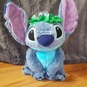 Puede incluir: Un peluche de Stitch azul con orejas grandes, una nariz azul y ojos negros grandes. La muñeca tiene una corona floral verde y está sentada sobre una superficie de madera. El peluche es un personaje popular de una película de Disney.