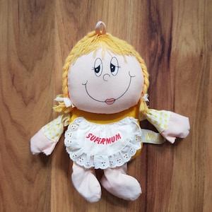 Peut inclure: Poupée vintage Supermum avec un visage souriant, des cheveux blonds tressés et une robe jaune avec des accents roses et blancs. La poupée porte un bavoir blanc avec le mot "Supermum" en lettres rouges.