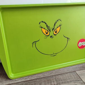 Könnte beinhalten: Ein rechteckiges, grünes Serviertablett mit dem Gesicht des Grinch, gelben Augen und einem verschmitzten Grinsen. Auf der rechten Seite befindet sich ein roter runder Aufkleber mit dem Wort "Grinch". Das Tablett hat abgerundete Kanten.