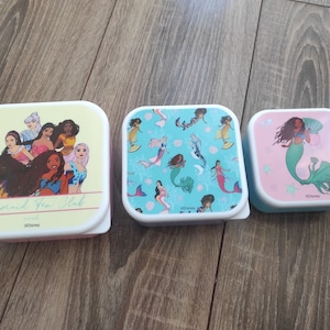 Puede incluir: Tres cajas de almuerzo cuadradas con un tema de sirena. La primera caja es amarilla con un grupo de sirenas y el texto "Mermaid Fan Club". La segunda caja es azul con un patrón de sirenas y conchas marinas. La tercera caja es rosa con una sola sirena con cabello largo verde y una concha.