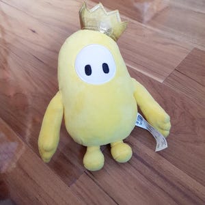 以下が含まれることがあります： 金色の王冠をかぶった、豆のようなキャラクターの形をした黄色のぬいぐるみ。おもちゃは、2つの黒い楕円形の目と小さな腕と脚を持つ白い楕円形の顔をしています。木製の表面に配置されています。