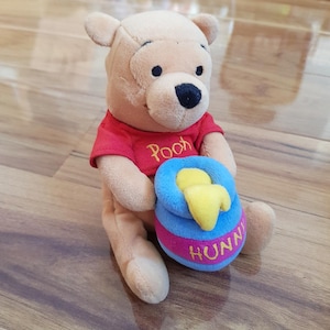 Könnte beinhalten: Ein Plüsch-Winnie-the-Pooh-Bär, der ein rotes Hemd trägt und einen blauen Honigtopf mit einem gelben Honiglöffel hält. Der Honigtopf hat die Aufschrift "Hunny" auf der Seite.