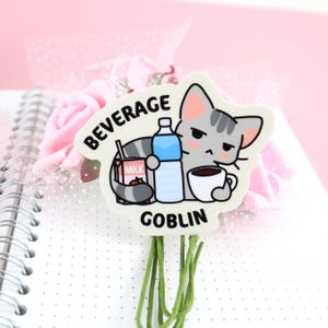 May include: A white sticker with a cartoon gray cat holding a cup of coffee, a carton of milk, and a bottle of water. The text "BEVERAGE GOBLIN" is printed on the sticker.