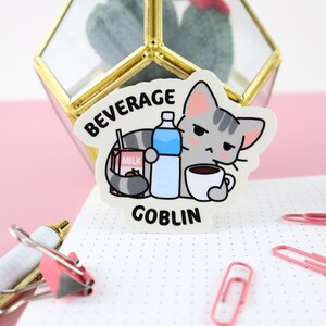 May include: A white sticker with a cartoon gray cat holding a carton of milk, a bottle of water, and a cup of coffee. The sticker says "Beverage Goblin" in black text.