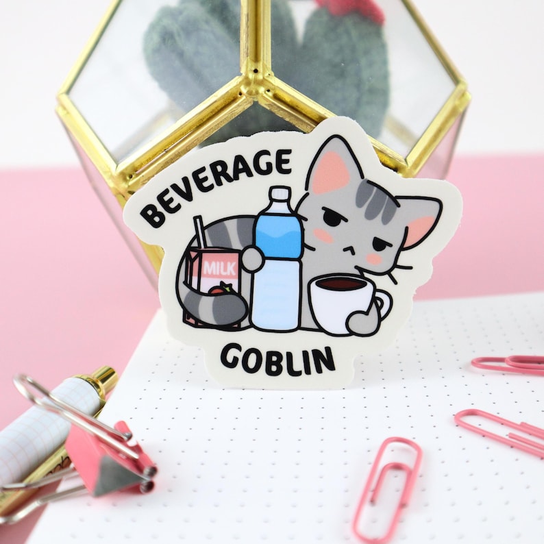 May include: A white sticker with a cartoon gray cat holding a carton of milk, a bottle of water, and a cup of coffee. The sticker says "Beverage Goblin" in black text.