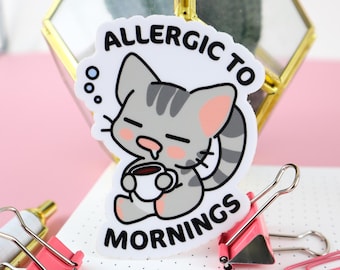 Op de afbeelding: Witte sticker met een cartoon grijze kat die een kopje koffie vasthoudt. De kat heeft een slaperige uitdrukking en de tekst "Allergic to Mornings" staat rond de kat.