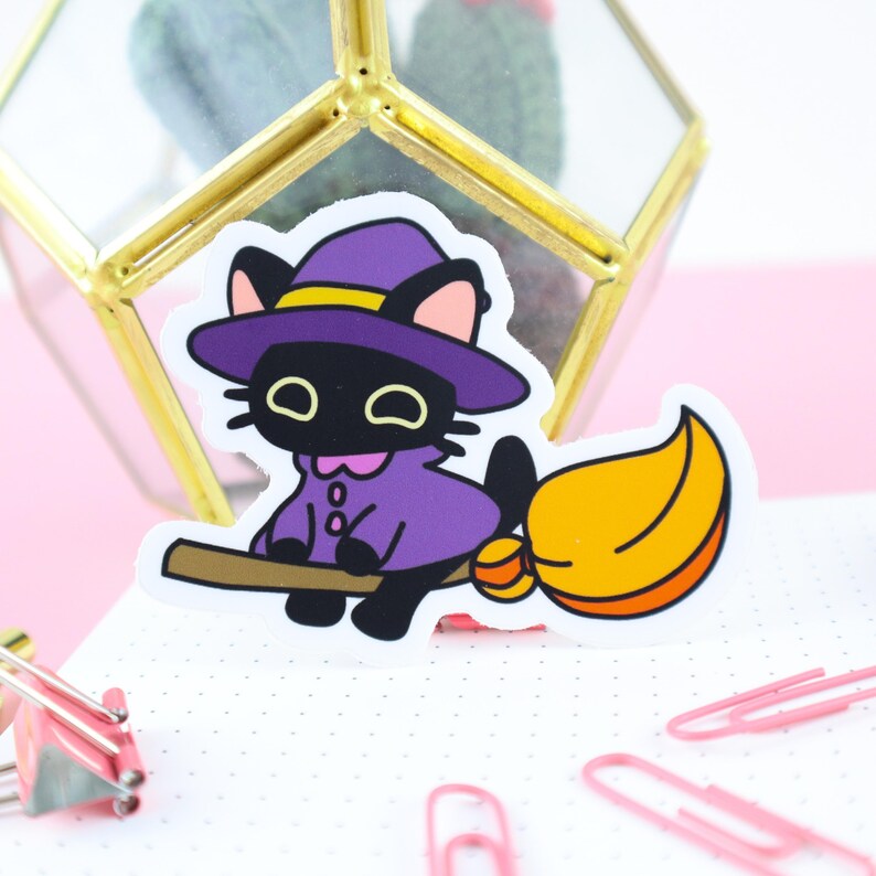 Pu&ograve; includere: Un adesivo raffigurante un gatto nero con un cappello e un mantello da strega viola, in sella a una scopa. Il gatto ha occhi gialli e orecchie rosa. La scopa &egrave; marrone con una spazzola arancione e gialla. L'adesivo &egrave; su una superficie bianca a pois.