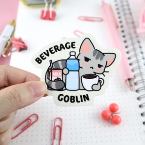 May include: A white sticker with a cartoon gray cat holding a cup of coffee, a carton of milk, and a bottle of water. The text "BEVERAGE GOBLIN" is printed on the sticker.