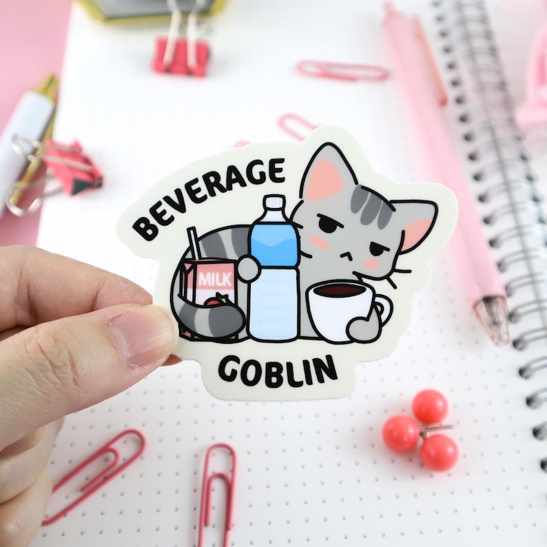 May include: A white sticker with a cartoon gray cat holding a cup of coffee, a carton of milk, and a bottle of water. The text "BEVERAGE GOBLIN" is printed on the sticker.