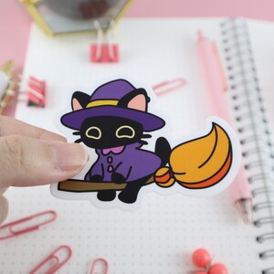 Pu&ograve; includere: Un adesivo con un gatto nero con un cappello e un mantello da strega viola, in sella a una scopa. Il gatto ha gli occhi gialli e sorride. La scopa ha una spazzola arancione e gialla. L'adesivo &egrave; su sfondo bianco con accenti rosa.
