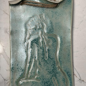 Puede incluir: Colgante de pared rectangular de cerámica con esmalte turquesa y superficie texturizada. La obra de arte presenta un relieve en relieve de una figura sentada. Se adjunta un lazo de cuerda natural para colgar.