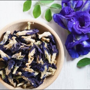 K&ouml;nnte beinhalten: Eine Holzschale gef&uuml;llt mit getrockneten blauen Schmetterlingserbsenbl&uuml;ten. Frische blaue Bl&uuml;ten und gr&uuml;ne Bl&auml;tter sind auf einer wei&szlig;en Oberfl&auml;che angeordnet. Das Logo "MAI'S FAMILY" befindet sich in der oberen linken Ecke.