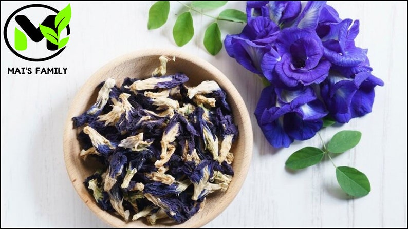 K&ouml;nnte beinhalten: Eine Holzschale gef&uuml;llt mit getrockneten blauen Schmetterlingserbsenbl&uuml;ten. Frische blaue Bl&uuml;ten und gr&uuml;ne Bl&auml;tter sind auf einer wei&szlig;en Oberfl&auml;che angeordnet. Das Logo "MAI'S FAMILY" befindet sich in der oberen linken Ecke.