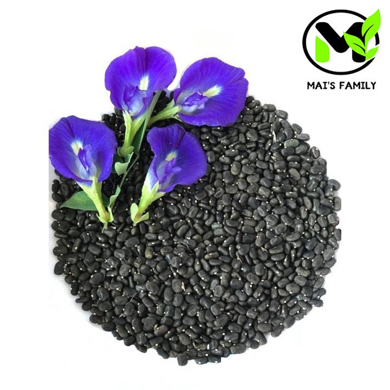 K&ouml;nnte beinhalten: Nahaufnahme von schwarzen Samen mit drei leuchtend violetten Schmetterlingsbl&uuml;ten. Die Bl&uuml;ten haben ein zartes Aussehen mit einem wei&szlig;en Zentrum. Die Samen sind verstreut und bilden eine strukturierte Oberfl&auml;che. Der Text "MAI'S FAMILY" ist in einem kreisf&ouml;rmigen Logo enthalten.